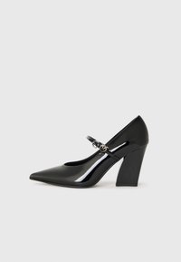 Chaussure à talon haut en cuir verni noir avec un bout pointu, un talon épais et une sangle avec des détails en métal.