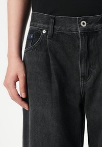 Čierne džínsy so slim fit strihom, s tradičným piatym vreckom a modrým šitím na prednom vrecku. Strieborné kovové akcenty.