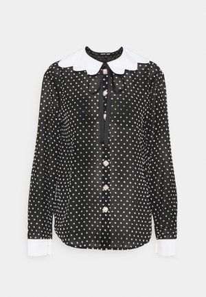 Blouse noire à manches longues avec des pois blancs, grand col blanc festonné, poignets blancs, boutons sur le devant, et un ruban noir noué au niveau du col.
