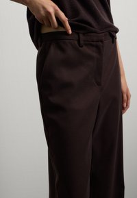 Persoon in donkerbruine broek en bijpassend topje, die het shirt een beetje oplicht en naar de taille wijst tegen een effen lichte achtergrond.