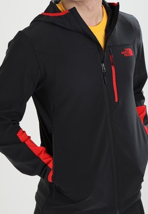 Homme portant une veste softshell noire avec des accents rouges et un logo, zippée à mi-hauteur, mains dans les poches, sur une chemise jaune.