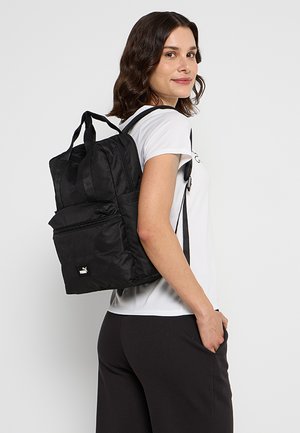 Kobieta w białej koszulce i czarnych spodniach niesie czarny plecak Puma na ramieniu, patrzy za siebie i się uśmiecha.