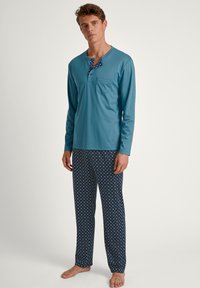 Turquoise langwerpige top met een zak en knoopsluiting; gecombineerd met navy broek met een wit vierkant patroon. Zachte stof.