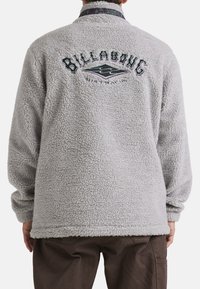 Grå sherpa fleece jakke med høj krave, struktureret materiale og "BILLABONG" broderet på ryggen i kontrasterende farver.