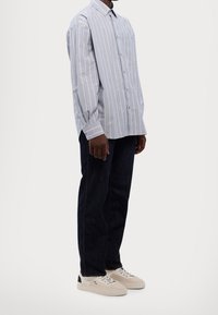 Camisa de botones a rayas en azul claro y blanco, de manga larga, con un solo bolsillo en el pecho; llevada con jeans de mezclilla oscuros y zapatillas beige.