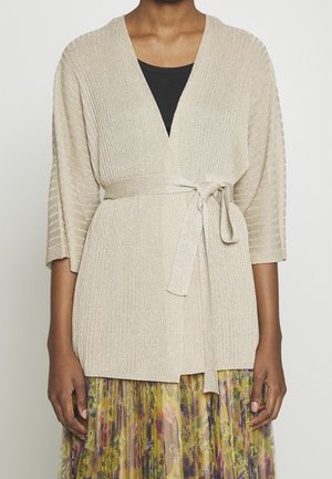 Femme portant un cardigan cache-cœur côtelé beige noué à la taille, un haut noir en dessous, et une jupe plissée fleurie aux tons jaunes et violets.