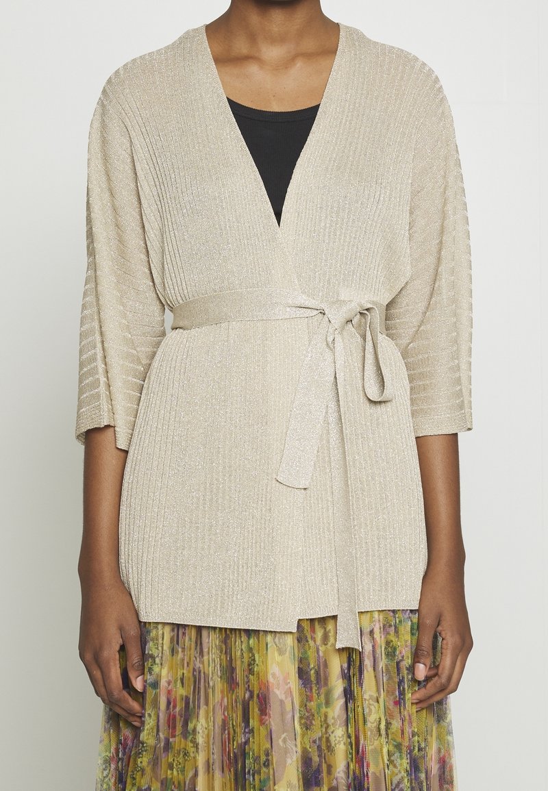 Femme portant un cardigan cache-cœur côtelé beige noué à la taille, un haut noir en dessous, et une jupe plissée fleurie aux tons jaunes et violets.