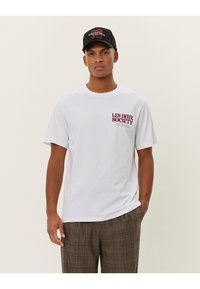 White cotton t-shirt featuring red printed "LES DEUX SOCIETY" logo on the left chest, paired with a black hat. Model standing against a light background.
