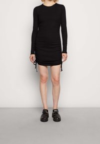 Svart långärmad klänning med rund urringning, med rynkade sidor med dragsko-detaljer och mjukt, stretchigt tyg. Bärs med svarta sandaler.