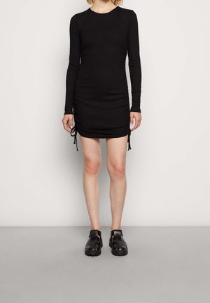 Schwarzes Langarmkleid mit rundem Ausschnitt, das an den Seiten geraffte Details mit Kordelzug und einen glatten, dehnbaren Stoff aufweist. Kombiniert mit schwarzen Sandalen.