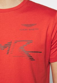 Czerwona koszulka bawełniana z czarnym logo Aston Martin i grafiką inspirowaną wyścigami. Krótkie rękawy i ribbowe wykończenie dekoltu.