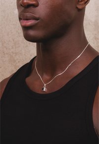 Silberne Kettenhalskette mit einem zylindrischen Anhänger und einem schwarzen Akzent, platziert auf einem schwarzen Tanktop. Einfaches, schlankes Design.