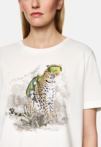 Weißes Baumwoll-T-Shirt mit einem Leoparden-Graphic, der grüne Sonnenbrillen und einen Rucksack trägt, umgeben von floralen Akzenten und einer strukturierten Bergkulisse.