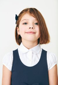 Mädchen in einem marineblauen Kleid mit rundem Ausschnitt über einem weißen Hemd mit Kragen; Haare mit einer blauen Schleife gestylt; neutraler Hintergrund.
