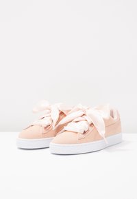 Helle rosa Wildleder-Sneaker mit großen Satinbändern als Schnürsenkel, weißer Gummisohle, glatter Textur und dezentem seitlichen Branding-Detail.