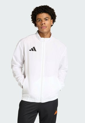 Junger Mann mit lockigem Haar, der eine weiße Jacke mit Reißverschluss und schwarzem Logo sowie schwarze Hosen mit orangefarbenem Logo trägt, Hände in den Taschen.