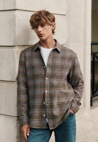 Bruine geruite overhemd met blauwe en oranje accenten, met knoopsluiting, lange mouwen en een gestructureerde stof; gedragen over een wit T-shirt.