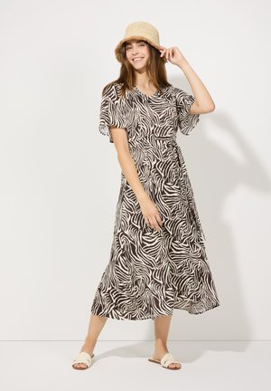 Robe imprimée zèbre marron et crème avec des manches volantées, taille cintrée et jupe mi-longue. Associée à des sandales claires et un chapeau en paille.