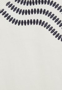 Fehér anyag, navy kék zigzag hímzéssel a felső részen, textúrált varrásmintával és sima felülettel.