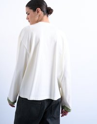 Γυναίκα με σκούρα μαλλιά πιασμένα κότσο, φορώντας λευκό πουκάμισο με μακριά μανίκια και πράσινα διακοσμημένα μανσέτα, καθώς και μαύρο τζιν, γυρισμένη από πίσω, μπροστά σε λευκό φόντο.