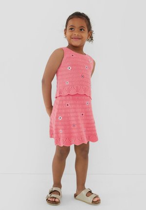 Jeune fille souriante, portant un top crochet sans manches rose et une jupe avec de petits motifs floraux ainsi que des sandales beiges.