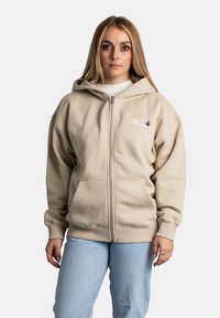 Sudadera beige con cremallera y corte relajado, grandes bolsillos frontales y un gráfico de mariposa en el lado izquierdo del pecho. Tejido suave de mezcla de algodón.