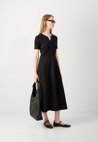 Black ribbed short-sleeve top with buttons, flowing black skirt, and black flats. Model holds a black leather tote bag.