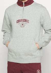 Szara polarowa bluza z ćwierć zamkiem w kolorze bordowym, z logo Converse i kieszenią typu kangur. Ściągacze przy mankietach i na dole.