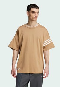 Brun oversized t-shirt i mjukt material, med tre vita ränder på ena ärmen och en liten logotyp på fållen. Korta, vida ärmar.