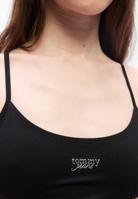 Jeune femme portant un haut noir à bretelles fines avec le logo "tommy jeans" brodé en fil argenté sur la poitrine.