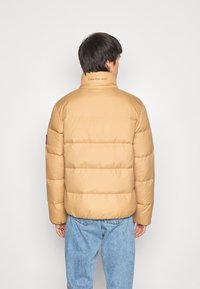 Tan pufferjakke med høj krave, der har quiltede sektioner, mærketekst på kraven og elastiske manchetter; båret over lyseblå jeans.