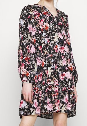 Femme portant une robe à manches longues, longueur genou, avec une base noire et un motif floral multicolore rose, rouge et blanc.