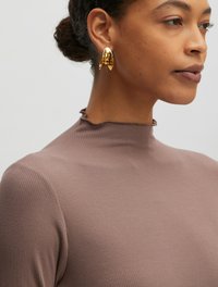 Brauner, gerippter Rollkragenpullover mit einem ruchengesäumten Ausschnitt, kombiniert mit glänzend goldenen, abstrakten Ohrringen. Der Stoff hat ein weiches, strukturiertes Aussehen.