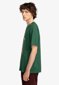 Camiseta de algodón verde con cuello redondo y mangas cortas, que presenta una impresión gráfica en la parte frontal. La tela parece suave y tiene un corte holgado.