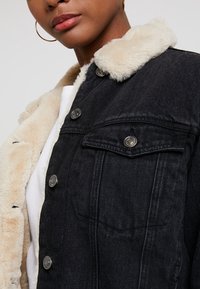 Svart denimjacka med beige faux fur-krage, silverknappar och framfickor. Nära bild som visar tygtexturen och design detaljer.