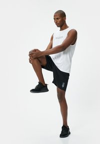 Top atlético sin mangas blanco con texto, pantalones cortos negros y zapatillas negras. El conjunto presenta un diseño ligero y un corte relajado.