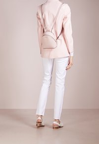 Malý pastelově růžový batoh s zakřiveným tvarem, hladkou texturou a minimalistickým designem. Obsahuje zlaté kovové doplňky a tenké nastavitelné popruhy.