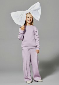 Sweatshirt et sweatpants en lavande en tissu doux. Grand nœud blanc maintenu au-dessus d'un sujet souriant, avec des cheveux lâches et des chaussures neutres.
