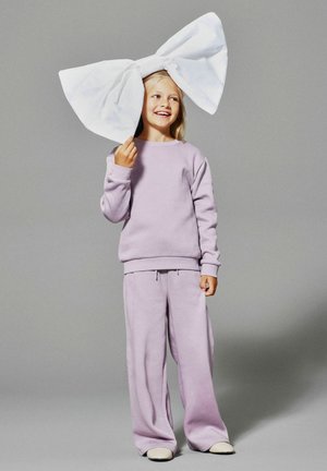 Felpa e pantaloni della tuta in lavanda realizzati in tessuto morbido. Grande fiocco bianco tenuto sopra un soggetto sorridente, con capelli sciolti e calzature neutre.