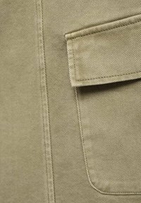 Tissu sergé beige avec un gros plan sur une poche rectangulaire dotée d'un rabat et de coutures doubles sur un vêtement.