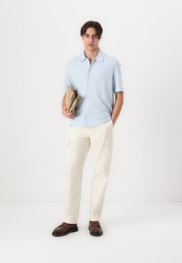 Camisa de botones de manga corta azul claro, pantalones cargo crema y mocasines marrones; sosteniendo un sobre beige en la mano izquierda; fondo blanco liso.