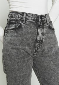 Jeans de mezclilla negros de tiro alto con un lavado desgastado, diseño de cinco bolsillos y detalles de costura visibles, que cuentan con un cierre delantero de botón y cremallera.