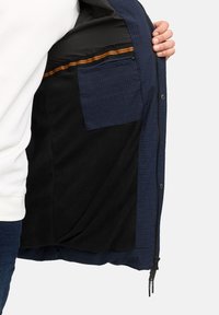 Navyjackets inre med svart fleecefoder, med en solid navyfärgad ficka och ett strukturerat mönster. Orange accentrand synlig.