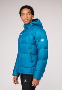 Blue puffer jacket with a hood, zippered front, and side pockets. Features quilted design and logo on sleeve. Worn over black pants.