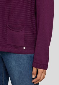 Lila gestrickter Pullover mit vertikaler Rippstruktur, einer Fronttasche und einem silbernen Logo-Akzent am Saum, kombiniert mit blauen Jeans.