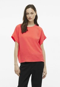 Top corallo a maniche corte con scollatura rotonda e tessuto testurizzato. Indossato con pantaloni neri, caratterizzati da una vestibilità rilassata e maniche a lunghezza media.