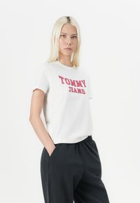 Camiseta de algodón blanca con "TOMMY JEANS" impreso en rojo, combinada con pantalones negros de corte holgado, con bolsillos laterales y cintura elástica.