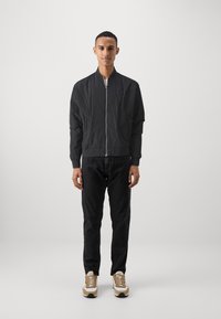 Veste bomber noire avec fermeture éclair, poignets côtelés et col montant. Portée sur un jean foncé et des chaussures de sport dans des tons neutres. Design minimaliste.