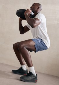 Fekete súlyzótányér, amelyet egy szürke sortot és fehér pólót viselő személy mindkét kezével tart, guggolást végezve. Fekete sportcipő és fehér zokni.