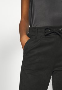 Pantalon en daim noir avec taille à cordon, poches latérales et texture lisse, assorti à un haut en satin gris, partiellement visible.